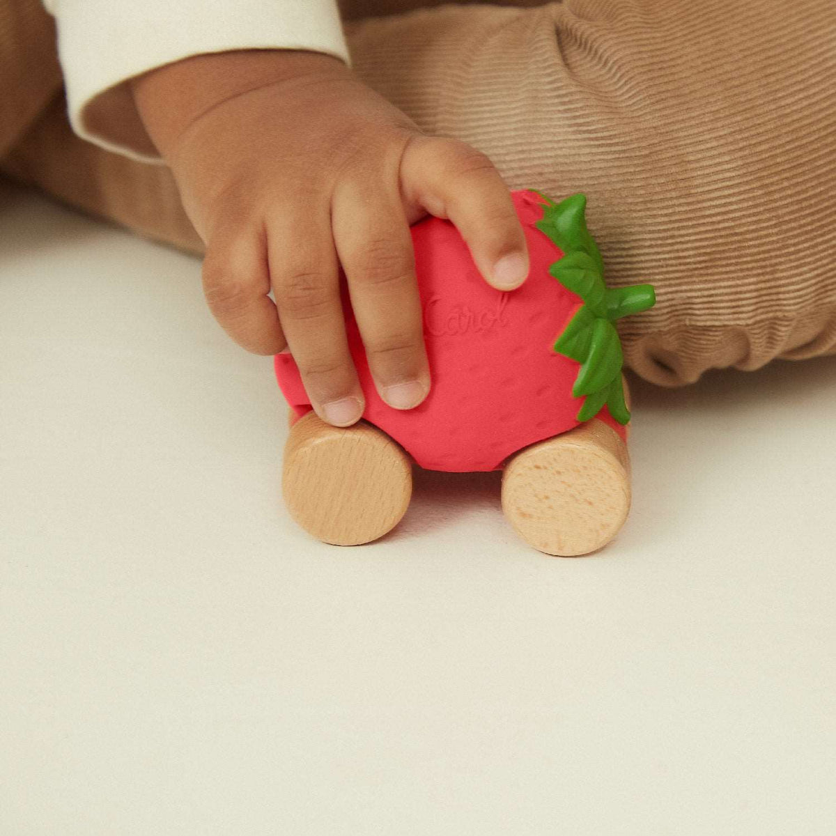 A toddler's hand holding a strawberry-shaped wooden toy car with a textured red body, green leafy top, and natural wood wheels.