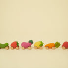 A line-up of various fruit and vegetable-shaped wooden toy cars with natural wood wheels, including a cucumber, strawberry, eggplant, lemon, carrot, and another strawberry on a neutral background.