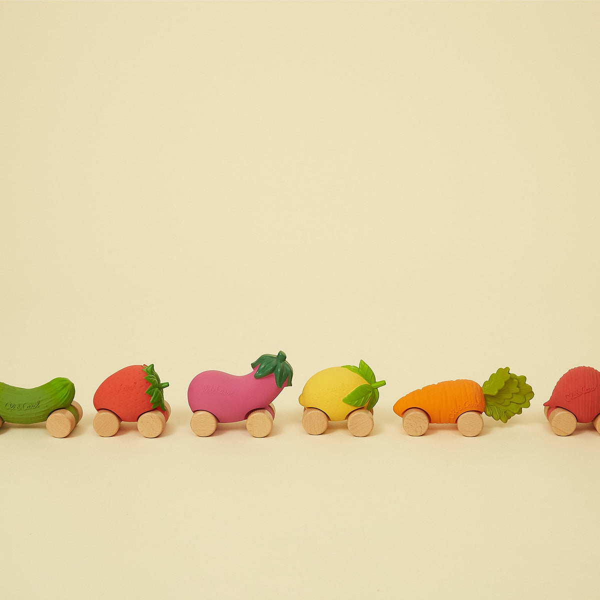 A line-up of various fruit and vegetable-shaped wooden toy cars with natural wood wheels, including a cucumber, strawberry, eggplant, lemon, carrot, and another strawberry on a neutral background.