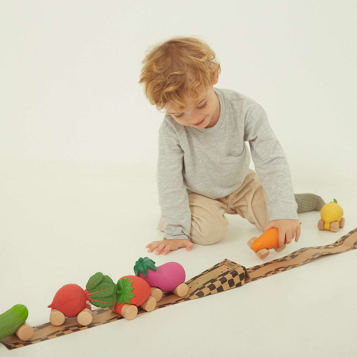A toddler playing on the floor with a checkered cardboard ramp and a line-up of various fruit and vegetable-shaped wooden toy cars.