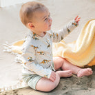 Baby sitting on sand wearing a dinosaur-patterned shirt and shorts