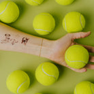 Hand holding a tennis ball with tattoos on arm surrounded by more tennis balls on a green background