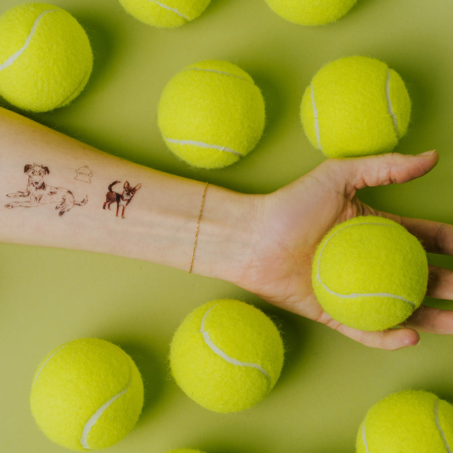 Hand holding a tennis ball with tattoos on arm surrounded by more tennis balls on a green background