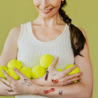 Woman holding tennis balls with visible tattoos on her arms against a green background