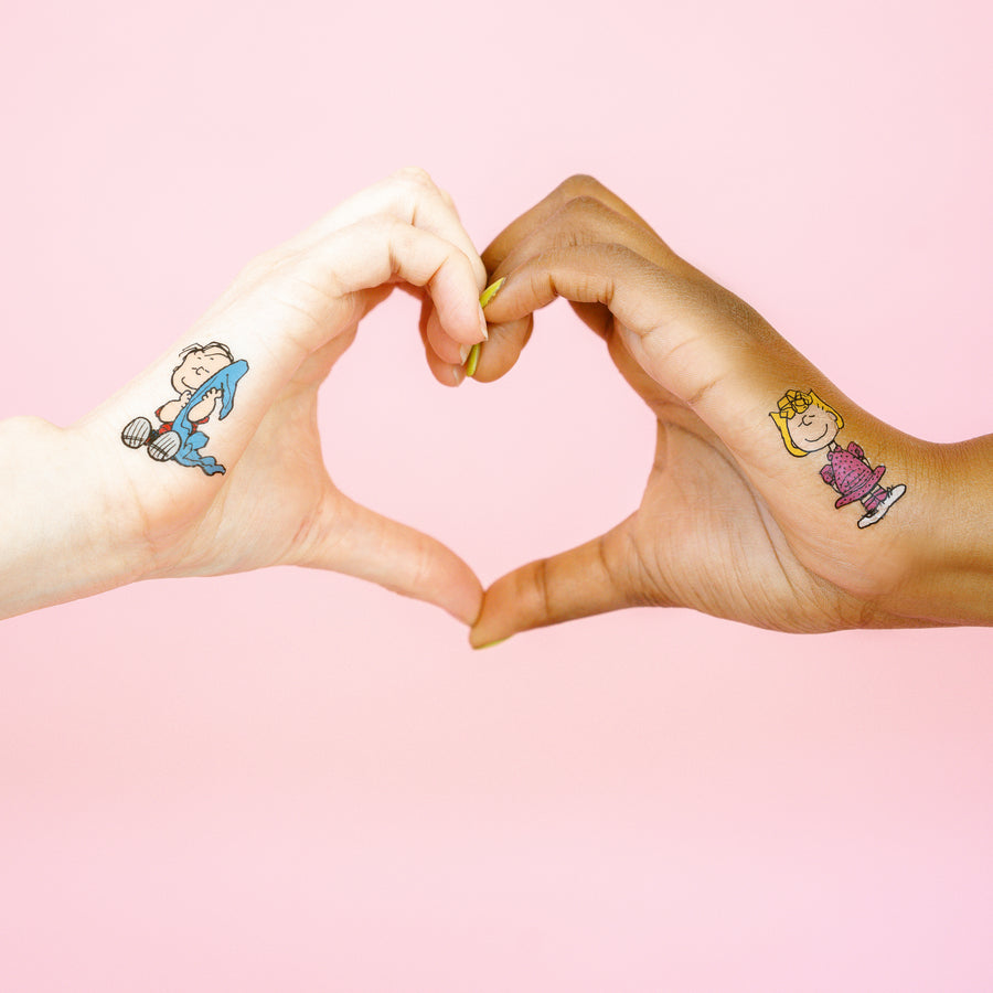 Two hands making a heart shape with cartoon character tattoos on a pink background