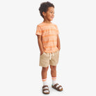 Child wearing a orange and white striped shirt with beige shorts on a white background