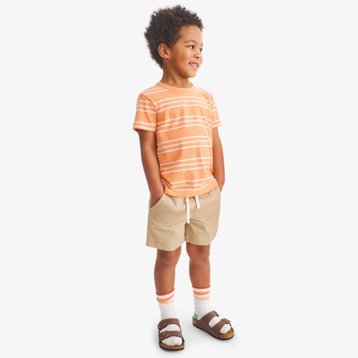 Child wearing a orange and white striped shirt with beige shorts on a white background