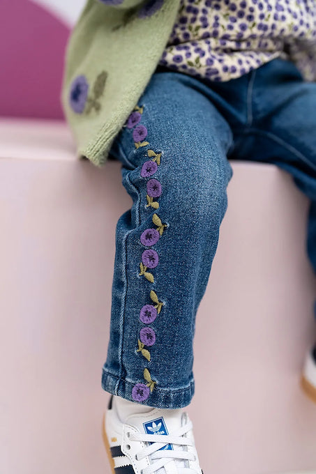 Close-up of a child wearing blue denim jeans with purple and yellow floral embroidery down the left leg, paired with white sneakers and a light green cardigan, seated on a pink bench.