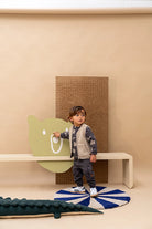 A toddler stands on a blue and white striped rug in a minimalist room, wearing a gray patterned cardigan, beige vest, and dark pants. The background features a beige wall, a wooden bench, a large green bear head cutout, and a brown textured panel.