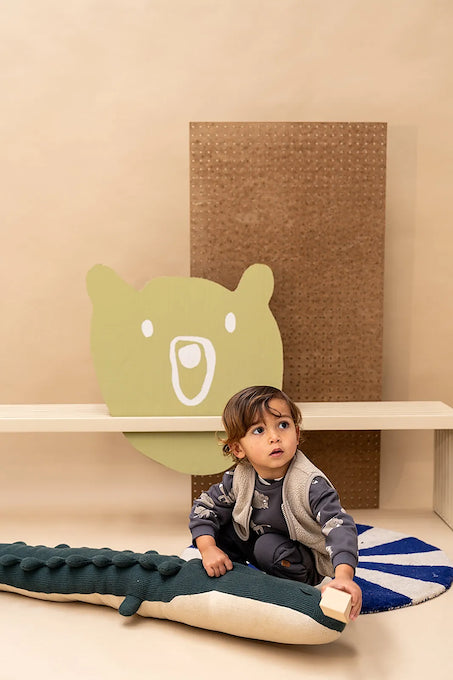 A toddler sits on a plush crocodile-shaped floor cushion in a styled nursery or playroom setting. The child is wearing a gray and white patterned long-sleeve shirt and a light gray vest.