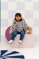 A young child sits on a pink block in a studio setting, wearing a patterned cream and navy jacket, a matching patterned top, blue jeans, and white sneakers. The background features a white and light purple checkered wall with a pink circular prop.