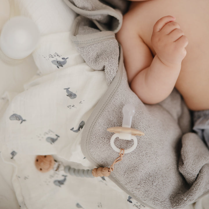 Baby wrapped in a towel with a pacifier, surrounded by soft toys on a neutral background
