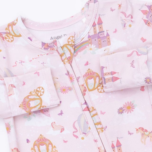 A close-up of a baby's pink zip-up sleeper or pajama top featuring a whimsical princess print with castles, carriages, unicorns, and flowers. The brand name 'Angel' is visible on the inner neck label.