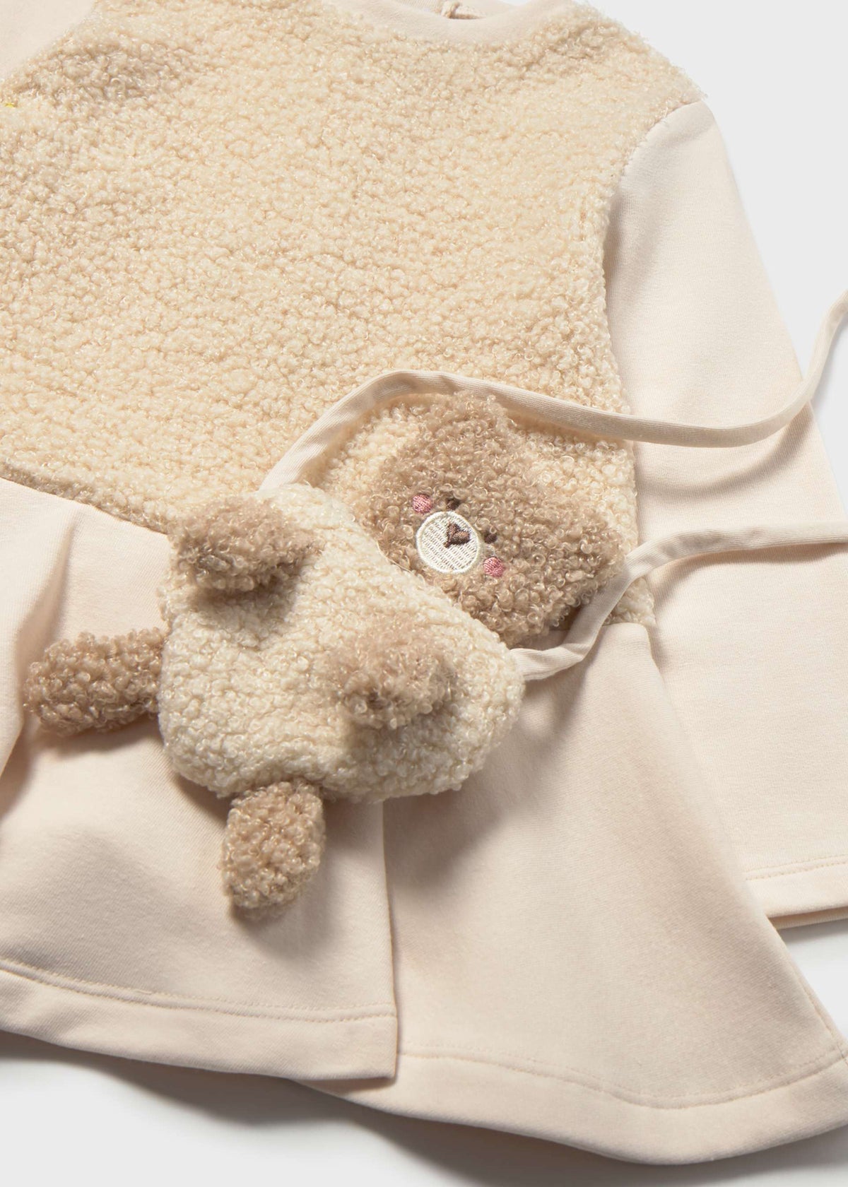 A cream-colored fleece teddy bear accessory with a small embroidered face, styled on top of a matching cream fleece garment.