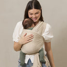 Woman holding a baby in a beige baby carrier against a plain background