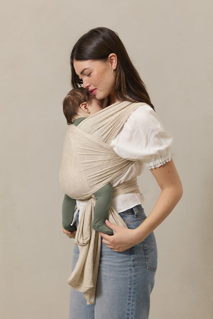 Woman holding a baby in a beige wrap against a plain background