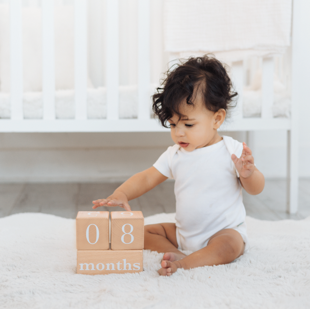 Baby sitting on a white blanket with wooden blocks indicating 8 months.