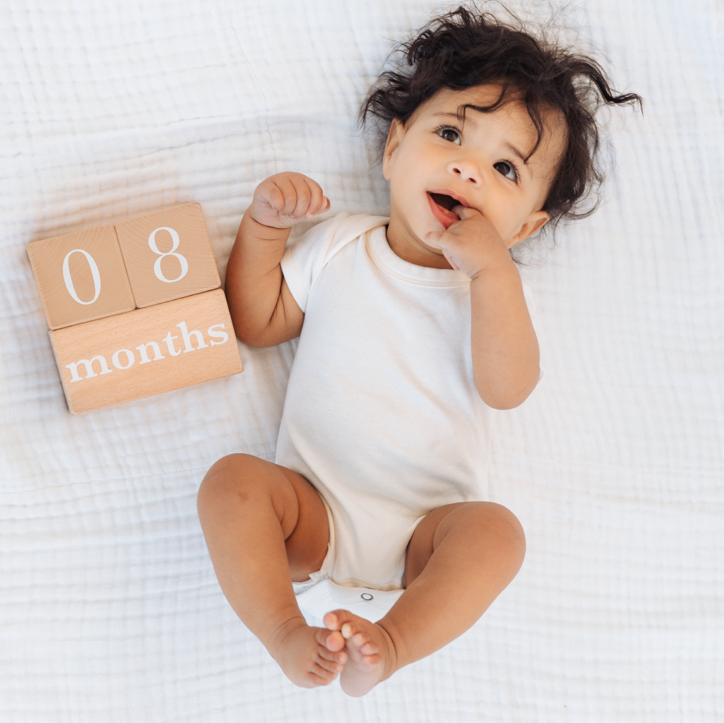 Baby in a white onesie sitting on a white blanket with blocks indicating 8 months.
