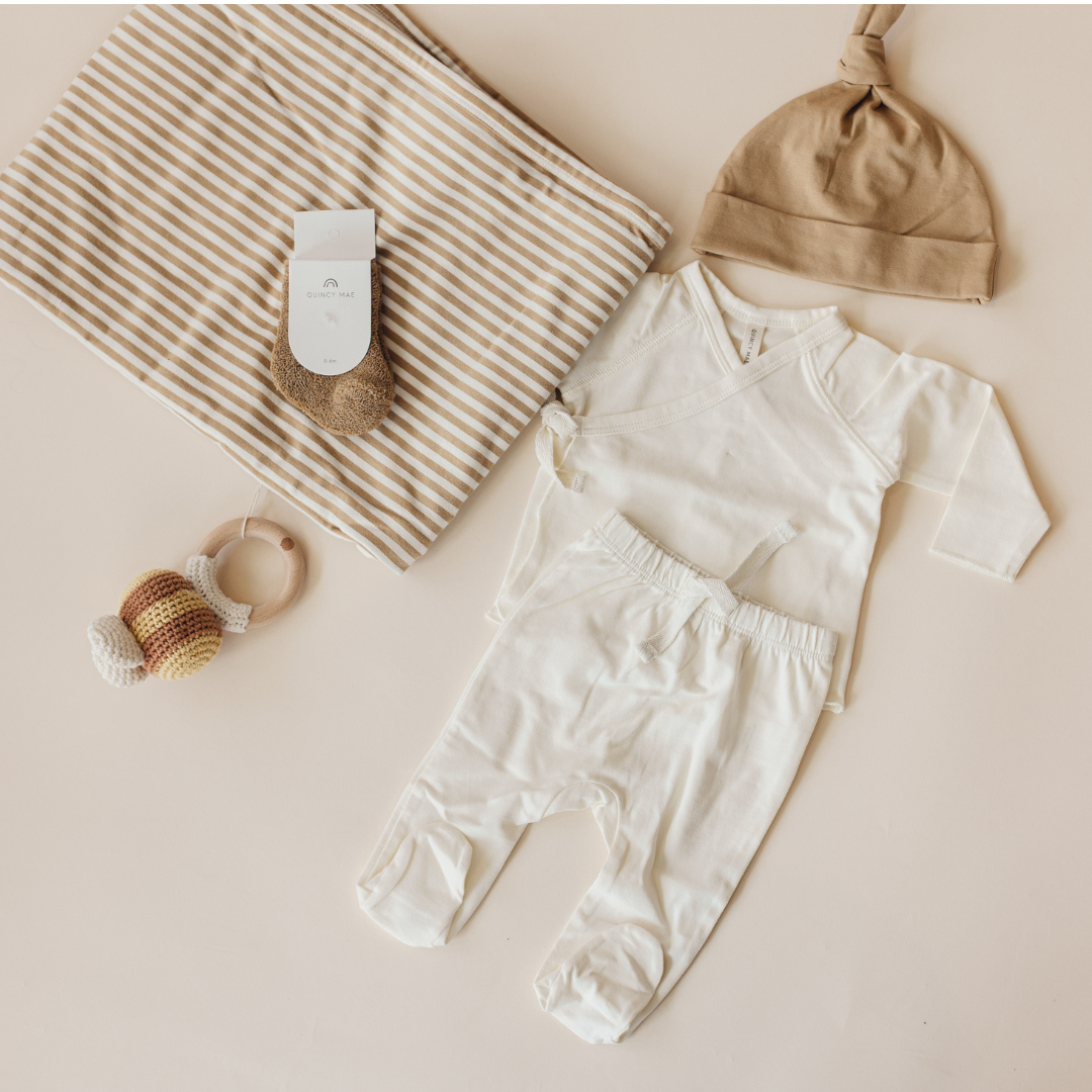 A baby clothing set laid flat on a warm beige background, including a white long-sleeve romper, matching white footed pants, and a tan knit beanie. A striped fabric item, a small knit bootie, and a wooden teething toy with a striped rattle are arranged nearby.