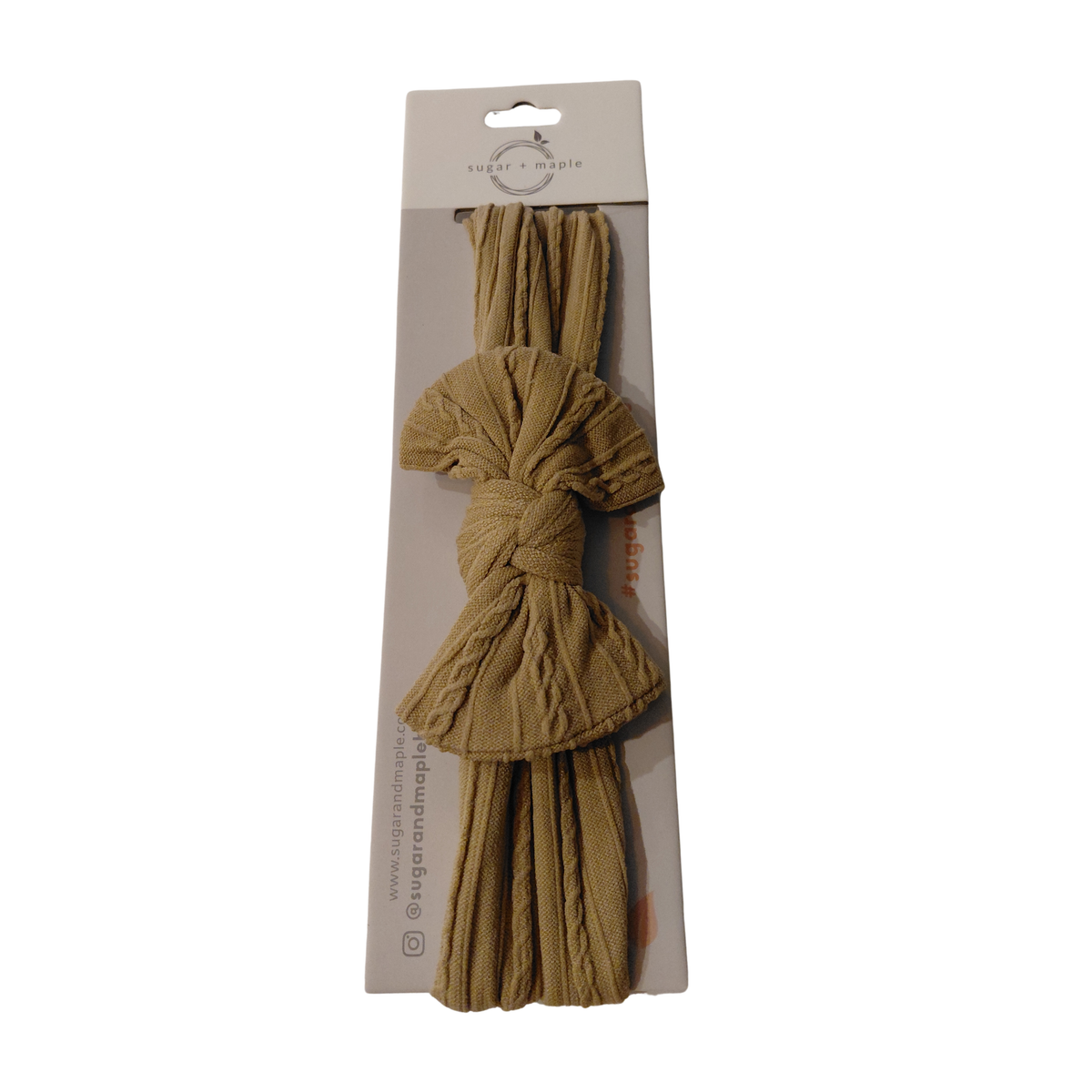 A packaged cotton tassel hair tie in a warm tan color, shown on a white background. The hair tie is knotted and displayed through a clear window in its original cardboard packaging.