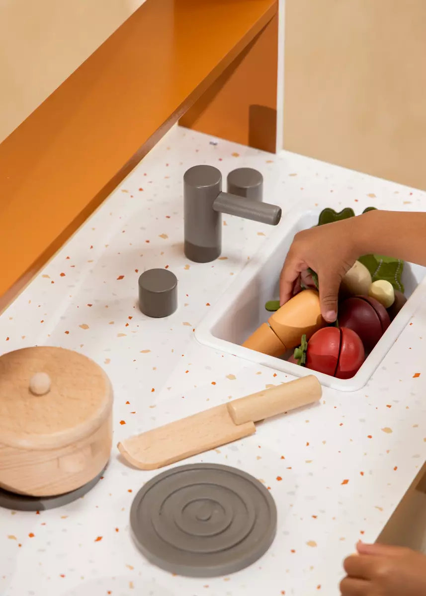 Children's play kitchen set with toy vegetables, sink, and accessories on a speckled countertop.