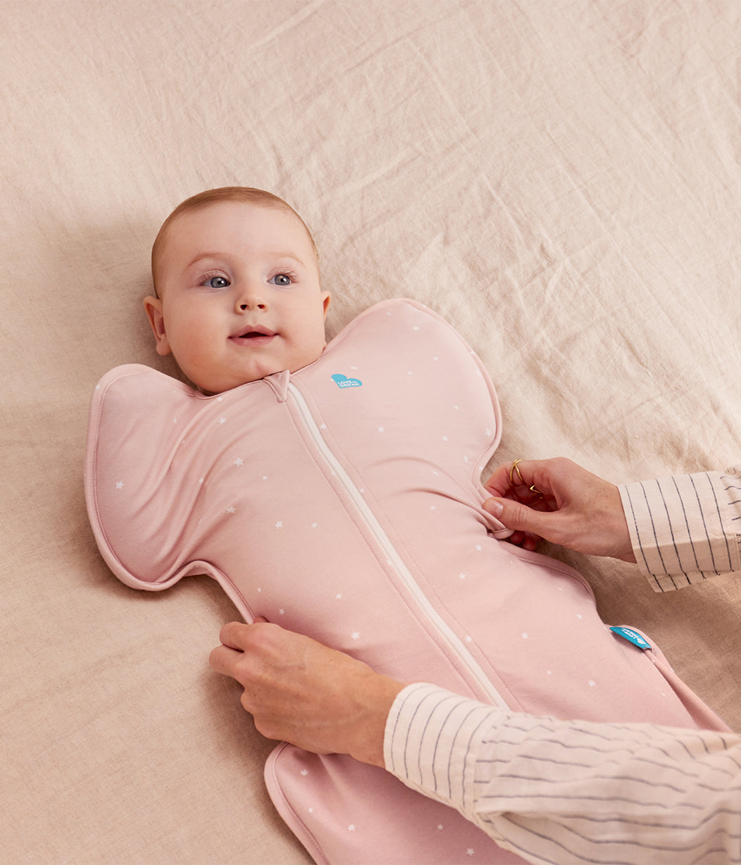 Baby lying on a pink blanket with a pink sleeping bag, being adjusted by an adult hand.