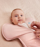 Baby in a pink sleeping bag with a person adjusting it on a soft surface