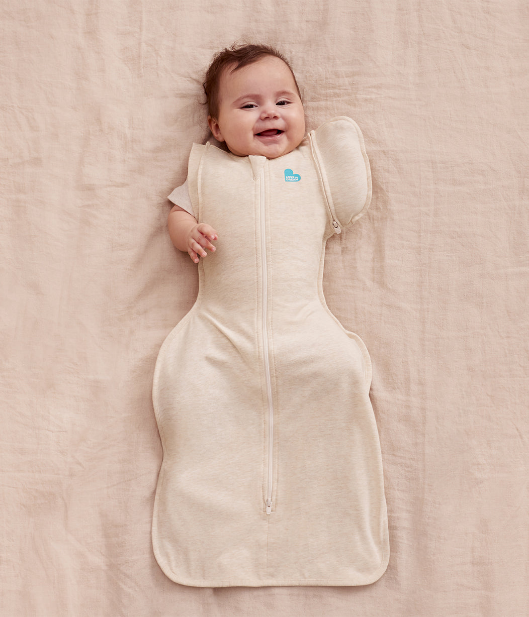 Baby lying on a beige blanket wearing a cream-colored sleep bag with a visible brand logo.