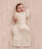 Baby lying on a beige blanket wearing a cream-colored sleep bag with a visible brand logo.