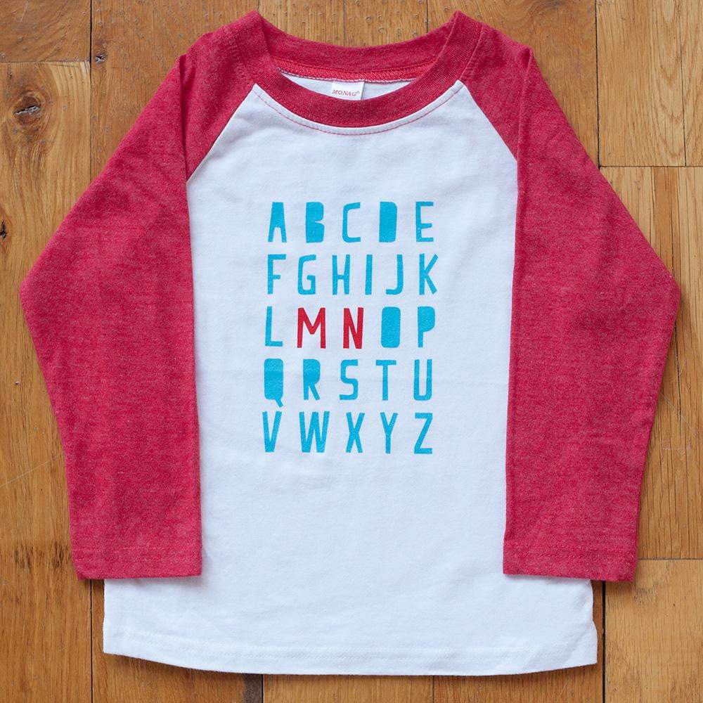 Red Minnesota alphabet raglan tee  on a wooden floor background.