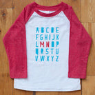 Red Minnesota alphabet raglan tee  on a wooden floor background.